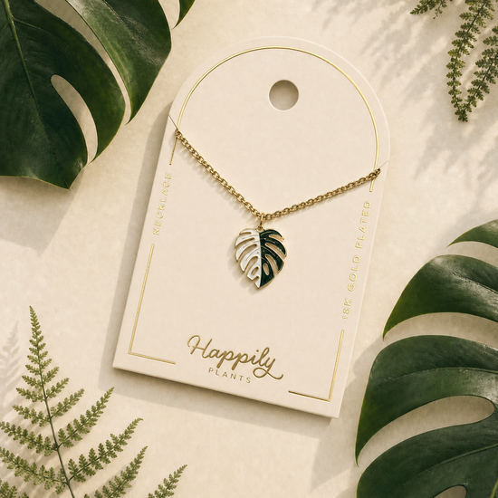 A gold leaf pendant necklace displayed on a cream-colored card with the brand name 'Happily Plants'. The card is styled in a flat-lay arrangement with green tropical leaves on a light beige background.
