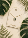 A gold leaf pendant necklace displayed on a cream-colored card with the brand name 'Happily Plants'. The card is styled in a flat-lay arrangement with green tropical leaves on a light beige background.
