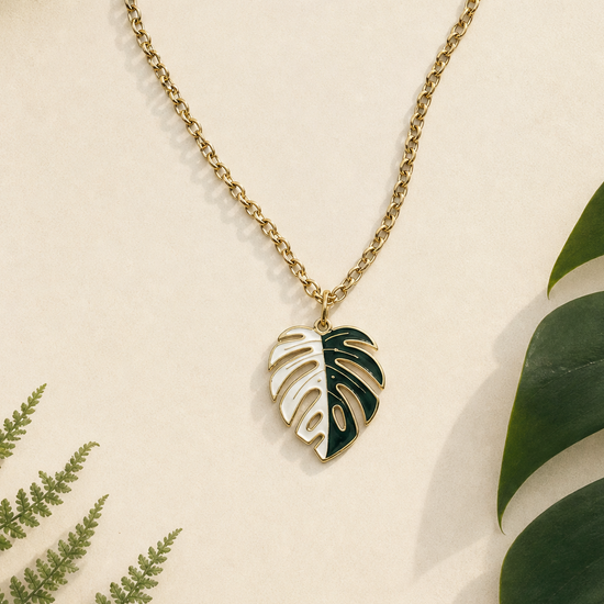 variegated monstera albo necklace A gold chain necklace with a split leaf pendant, half white and half green, styled on a cream background with tropical green leaves.